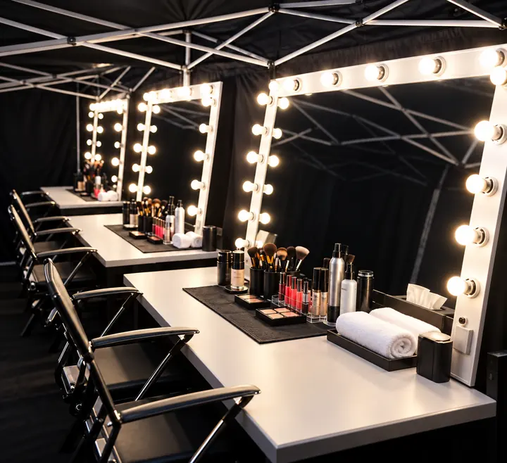 Estación de maquillaje bajo carpa negra con espejos iluminados y sillas plegables.